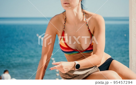 Woman in rainbow bikini. Happy tanned well looking middle aged woman in rainbow swimsuit at seaside, blue sea water in background. Holiday, vacation and recreational concept. 95457294