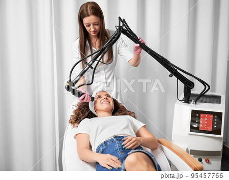 Visiting beautician to treat post-traumatic acne scars. Female dermatologist performing laser resurfacing on face skin of smiling young lady. Visiting beautician to treat post-traumatic acne scars. Female dermatologist performing laser resurfacing on face skin of smiling young lady. 95457766