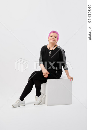 Happy old woman with short pink hair in long knitted dress sits on box 95459340