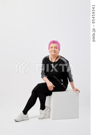 Happy old woman with short pink hair in long knitted dress sits on box leaning forward 95459341