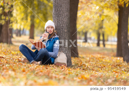 Digital detox. Adult woman in warm casual wear sits under a tree and reading a book drinking coffee in the autumn urban park outdoor, selected focus. 95459549