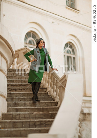 Outdoor fashion portrait of an elegant fashionable brunette model in a beret, green dress and a gray waistcoat posing at sunset in a european city in autumn. 95460235