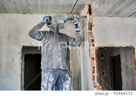 Strong repairman working with heavy special tool for destroying walls. Side view of builder with jackhammer during dismantling brick partition indoors. Strong repairman working with heavy special tool for destroying walls. Side view of builder with jackhammer during dismantling brick partition indoors. 95464133