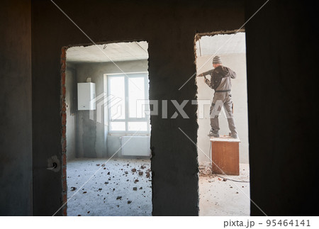 Man builder standing on wooden table and drilling wall between two rooms with doorways. Male worker using electric hammer drill while breaking the wall in apartment during renovation. Man builder standing on wooden table and drilling wall between two rooms with doorways. Male worker using electric hammer drill while breaking the wall in apartment during renovation. 95464141