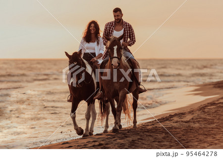 A loving couple in summer clothes riding a horse on a sandy beach at sunset. Sea and sunset in the background. Selective focus  95464278