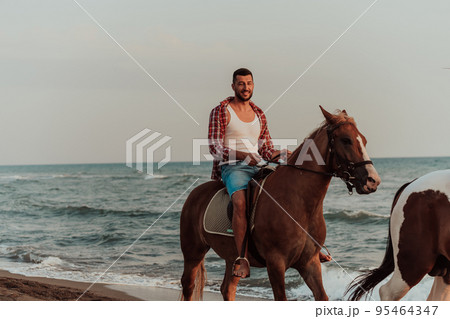 A loving couple in summer clothes riding a horse on a sandy beach at sunset. Sea and sunset in the background. Selective focus A loving couple in summer clothes riding a horse on a sandy beach at sunset. Sea and sunset in the background. Selective focus 95464347