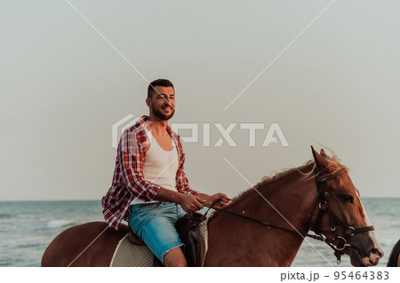 A loving couple in summer clothes riding a horse on a sandy beach at sunset. Sea and sunset in the background. Selective focus  95464383