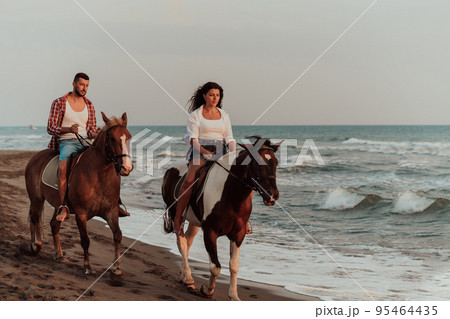 A loving couple in summer clothes riding a horse on a sandy beach at sunset. Sea and sunset in the background. Selective focus  95464435