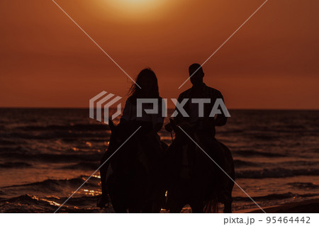 A loving couple in summer clothes riding a horse on a sandy beach at sunset. Sea and sunset in the background. Selective focus A loving couple in summer clothes riding a horse on a sandy beach at sunset. Sea and sunset in the background. Selective focus 95464442