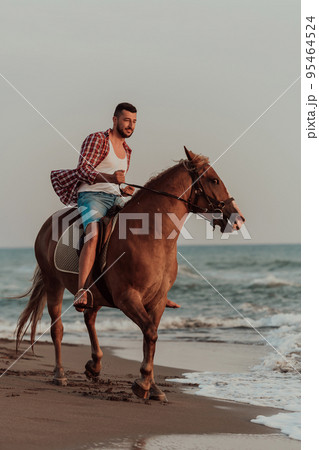 A modern man in summer clothes enjoys riding a horse on a beautiful sandy beach at sunset. Selective focus  95464524