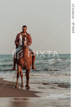 A modern man in summer clothes enjoys riding a horse on a beautiful sandy beach at sunset. Selective focus  95464525
