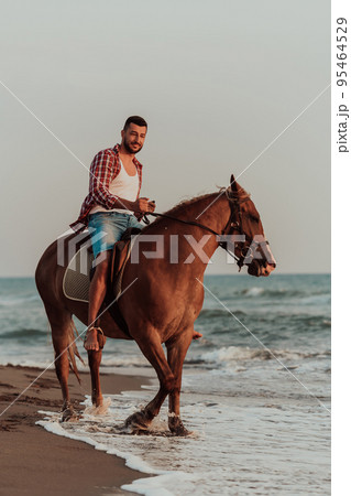 A modern man in summer clothes enjoys riding a horse on a beautiful sandy beach at sunset. Selective focus A modern man in summer clothes enjoys riding a horse on a beautiful sandy beach at sunset. Selective focus 95464529