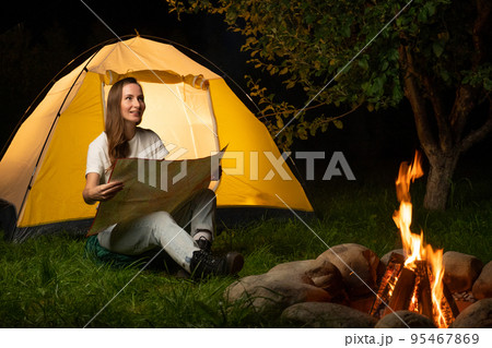Young woman is camping outdoors, sitting by a campfire and consulting a map against the background of a tourist tent Young woman is camping outdoors, sitting by a campfire and consulting a map against the background of a tourist tent 95467869