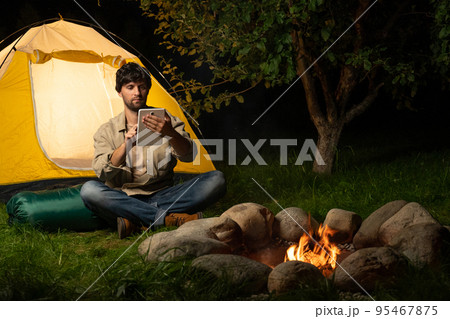 Happy tourist using a digital tablet while sitting by the campfire near his tent at night 95467875