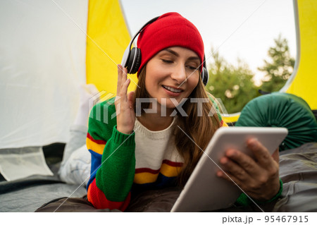 Beautiful woman in a red hat listens to music with headphones while sitting in a tourist tent 95467915
