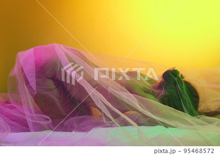 Portrait of tender woman covered in transparent fabric isolated over orange background in green pink neon light 95468572