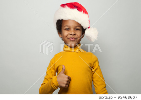 Small optimistic black child boy in Santa hat smiling and showing thumb up on white background Small optimistic black child boy in Santa hat smiling and showing thumb up on white background 95468607
