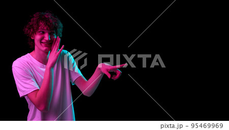 Portrait of young smiling man whispering isolated over dark background in purple neon light. Mental health, news, emotions, youth, sales, ad 95469969