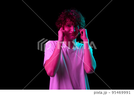 Terrible headache. Young man with discontent and undercurrent facial expression covering his ears isolated over dark background in neon light. 95469991
