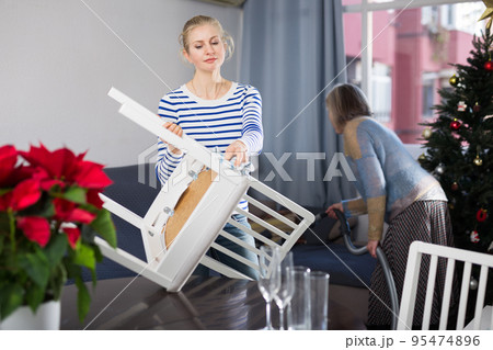 Young woman dusts chair while an elderly mother vacuums room before Christmas Young woman dusts chair while an elderly mother vacuums room before Christmas 95474896