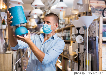 Man in protective mask is choosing new table lamp for her home in store Man in protective mask is choosing new table lamp for her home in store 95475013