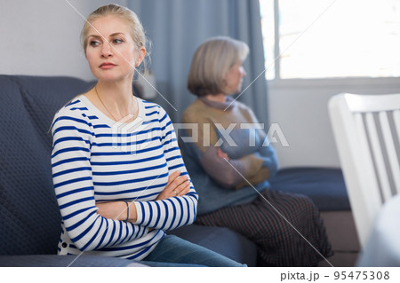 Elderly mother and adult daughter sit on the couch turned away from each other after domestic quarrel Elderly mother and adult daughter sit on the couch turned away from each other after domestic quarrel 95475308