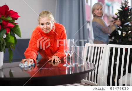 Adult daughter wipes a table with a cloth, while an elderly mother decorates Christmas tree Adult daughter wipes a table with a cloth, while an elderly mother decorates Christmas tree 95475339