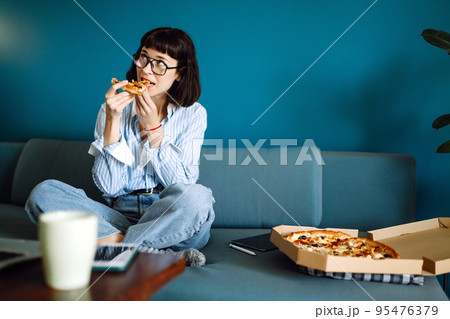 Young woman eating a piece of delicious pizza from the box, sitting at sofa at home. Food delivery. 95476379
