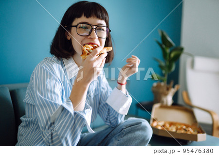 Young woman eating a piece of delicious pizza from the box, sitting at sofa at home. Food delivery. 95476380