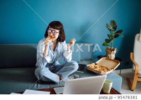 Young woman eating a piece of delicious pizza from the box, sitting at sofa at home. Food delivery. 95476381