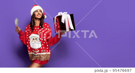 Beautiful girl in Santa's hat and christmas sweater holding glass of champagne and gift box in hands Beautiful girl in Santa's hat and christmas sweater holding glass of champagne and gift box in hands 95476697