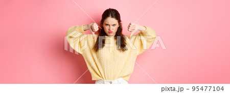 Angry and displeased young girl frowning, showing thumbs down at something bad, express dislike, leave negative feedback, standing upset against pink background Angry and displeased young girl frowning, showing thumbs down at something bad, express dislike, leave negative feedback, standing upset against pink background 95477104