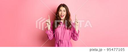 Beauty and fashion. Happy woman looking with amazement at copy space on top, pointing fingers up, smiling amazed, checking out promo offer, standing over pink background Beauty and fashion. Happy woman looking with amazement at copy space on top, pointing fingers up, smiling amazed, checking out promo offer, standing over pink background 95477105