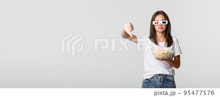 Disappointed attractive girl in 3d glasses, holding popcorn and showing thumbs-down, dislike movie 95477576