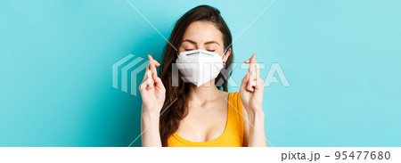 Health, covid-19 and lockdown concept. Close up of hopeful young woman in respirator mask, cross fingers and making wish, praying with eyes closed, standing against blue background 95477680