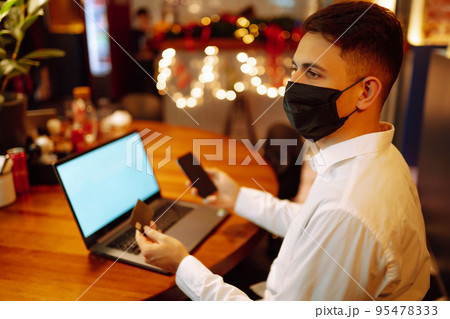 Young man wearing medical face mask working on laptop computer and speaks on the phone. Young man wearing medical face mask working on laptop computer and speaks on the phone. 95478333