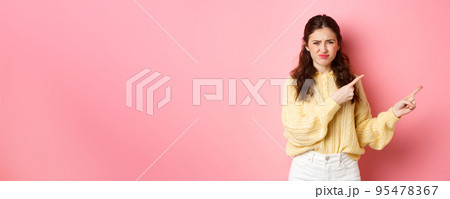 Skeptical and displeased young woman frowning, grimacing and pointing fingers right at copyspace, express dislike and disappointment, standing against pink background 95478367