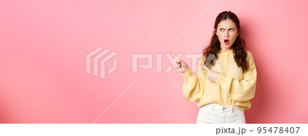 Offended and shocked young woman staring and pointing fingers left at copyspace aside, insulted by something, gasping displeased, standing against pink background Offended and shocked young woman staring and pointing fingers left at copyspace aside, insulted by something, gasping displeased, standing against pink background 95478407