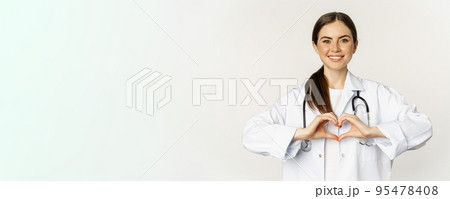 Portrait of smiling doctor, female physician showing heart sign and looking with care, love her patients, standing over white background Portrait of smiling doctor, female physician showing heart sign and looking with care, love her patients, standing over white background 95478408