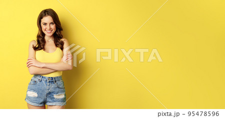Summer holidays and people. Stylish bright girl in tank top and shorts, posing against yellow background, smiling and looking relaxed Summer holidays and people. Stylish bright girl in tank top and shorts, posing against yellow background, smiling and looking relaxed 95478596