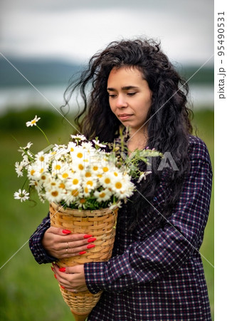 A woman stands on a green field and holds a basket with a large bouquet of daisies in her hands. In the background are mountains and a lake. A woman stands on a green field and holds a basket with a large bouquet of daisies in her hands. In the background are mountains and a lake. 95490531