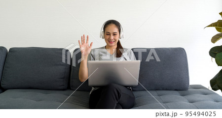 Asian woman in gray t-shirt and headphone waving hand via remote communication through laptop computer in the living room. Asian woman in gray t-shirt and headphone waving hand via remote communication through laptop computer in the living room. 95494025