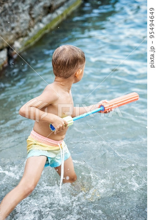 Small boy holds bright water-jet gun standing in sea Small boy holds bright water-jet gun standing in sea 95494649
