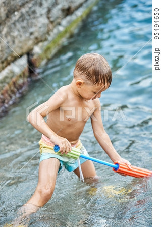 Small boy holds bright water-jet gun standing in sea 95494650
