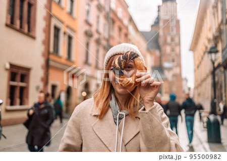 Happy young blonde woman tourist in stylish clothes holding pretzel obwarzanek on the market square in Krakow in Poland Happy young blonde woman tourist in stylish clothes holding pretzel obwarzanek on the market square in Krakow in Poland 95500982