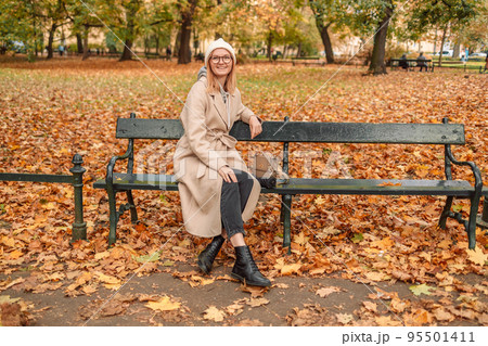 Autumn woman in a stylish beautiful warm coat from a knitted hat relaxes on a bench on a warm day 95501411
