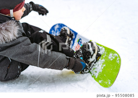 little boy sitting on snow putting his feet in snowboard bindings adjusting straps little boy sitting on snow putting his feet in snowboard bindings adjusting straps 95508085