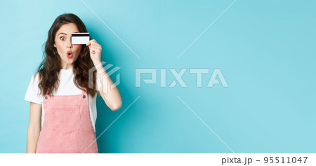 Excited girl holding plastic credit card near eye and look amazed at camera, buying things in store, going shopping, wasting money on bank account, standing over blue background 95511047