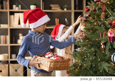Two sibling kids in red Santa hats decorating fir tree 95516088