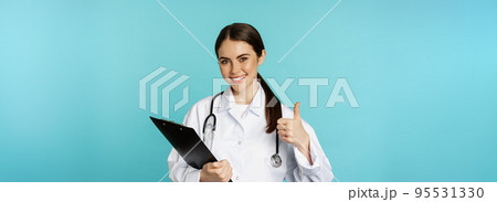 Confident smiling doctor woman, physician showing thumbs up, holding clipboard, appointment in hospital, standing over torquoise background 95531330